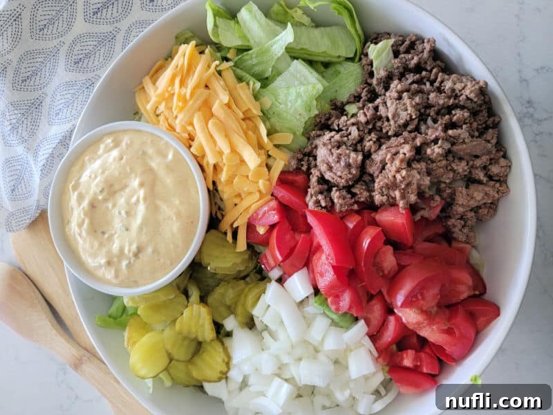 Big Mac Salad in a white bowl with big mac sauce, dill pickles, onions, tomatoes, ground beef, lettuce, and shredded cheddar cheese in a white bowl with wooden spoons and a cloth napkin