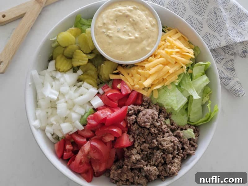 Big Mac Salad in a white bowl with big mac sauce, dill pickles, onions, tomatoes, ground beef, lettuce, and shredded cheddar cheese. 