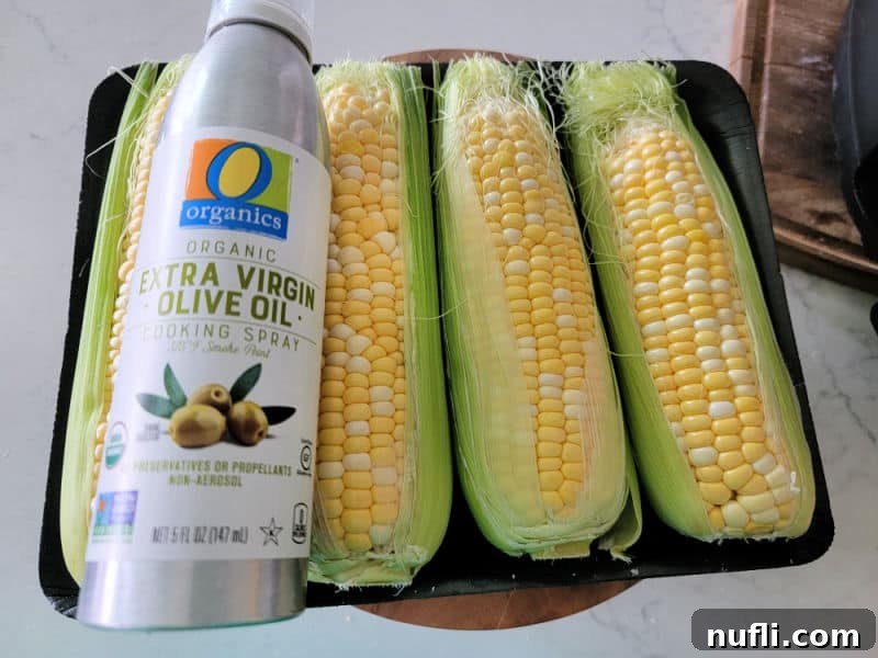 Olive oil cooking spray bottle next to two ears of fresh corn on the cob