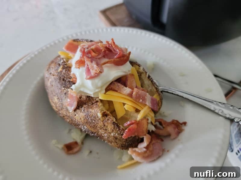 bacon, sour cream, cheese, on a baked potato on a white plate with a fork next to an air fryer 
