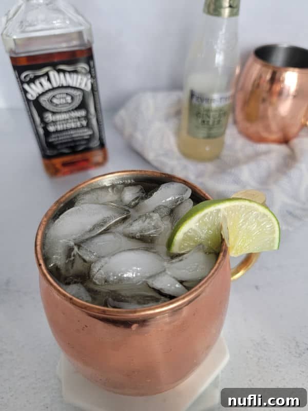 Copper mug with cocktail and lime slice in front of a Jack Daniels bottle, ginger beer bottle, and a 2nd copper mug