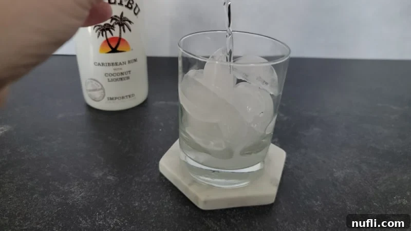 Clear liquid poured into a glass with ice cubes. Bottle of Malibu in the background.