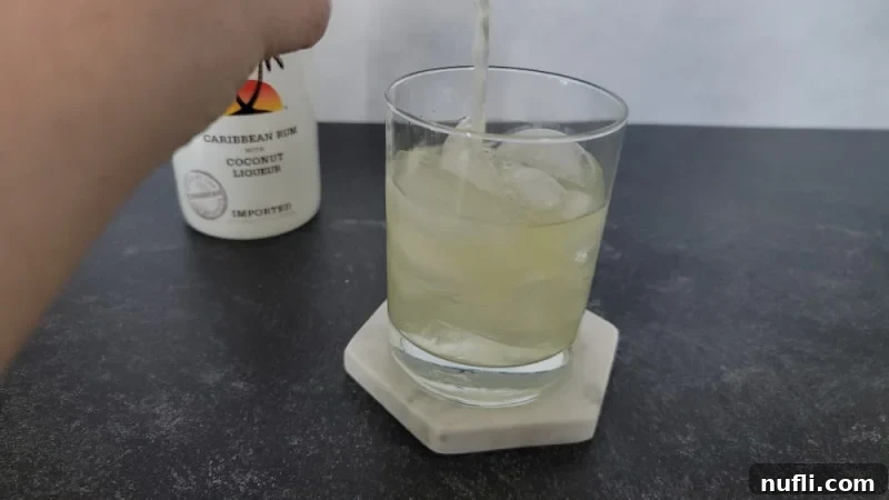 Pineapple juice pouring into a glass with ice and a bottle of Malibu in the background.