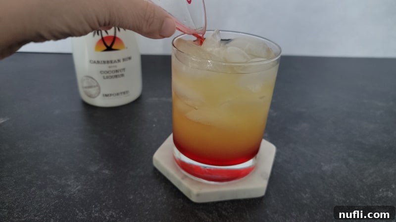 Red liquid pouring into a yellow liquid in a clear glass with a bottle of Malibu rum in the background.