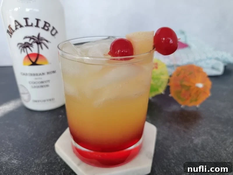 Red and yellow layered cocktail with maraschino cherry and pineapple chunk garnish in front of a bottle of Malibu coconut rum.