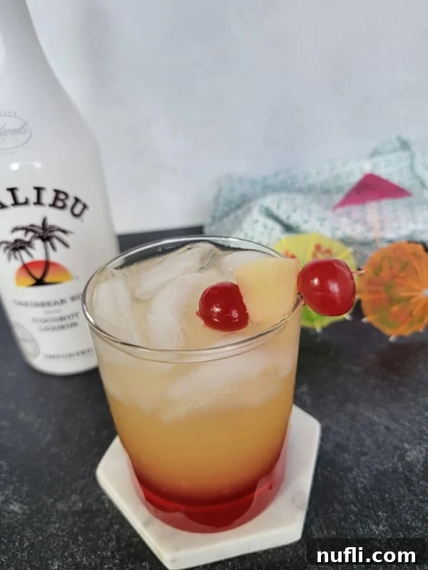 Layered yellow and red liquid in a clear glass with a garnish of maraschino cherries and pineapple chunks. Malibu coconut rum bottle in the background