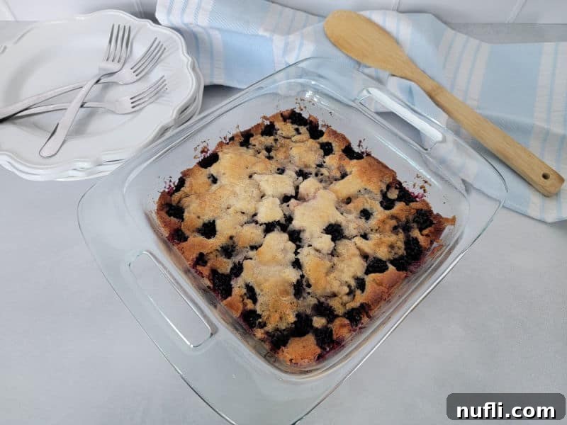 Bisquick Blackberry Cobbler in a glass baking dish next to a stack of plates and a wooden spoon on a cloth napkin, ready to be served.