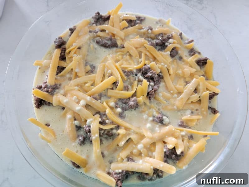 Bisquick Impossible Cheeseburger Pie with shredded cheddar cheese, ground beef, and bisquick mixture in a glass pie dish
