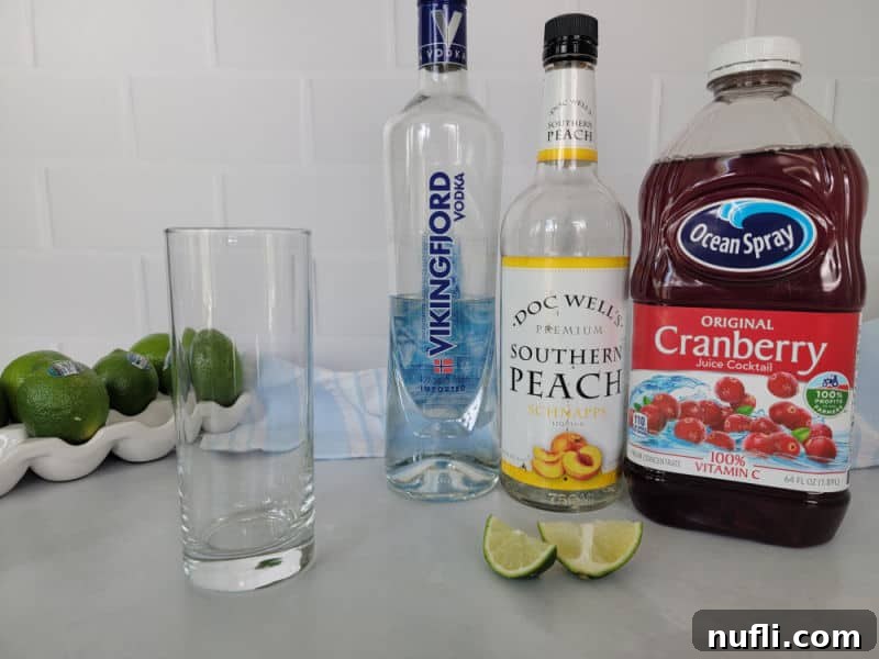 Effortless 3-Ingredient Woo Woo Cocktail 3 Woo Woo drink ingredients, fresh limes, vodka, peach schnapps, cranberry juice next to a tall old fashioned glass