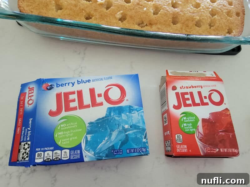 Red and blue Jell-O boxes displayed prominently in front of a glass baking dish containing a freshly baked white cake, ready for preparation.
