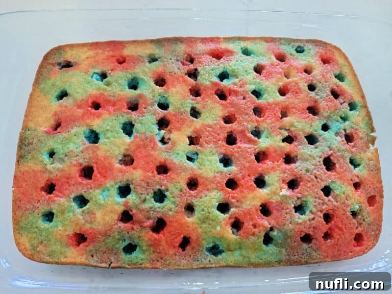 A close-up shot of a white cake baked in a glass dish, featuring visible holes filled with vibrant red and blue Jell-O, demonstrating the poke cake technique.