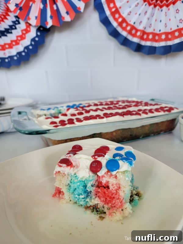 A perfect square slice of Red, White, and Blue Poke Cake on a white plate, positioned in front of the full cake in its baking dish and decorative American flags.