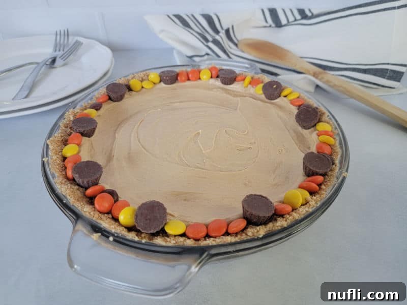 peanut butter pie rimmed with Reese's pieces in a glass baking dish on a counter with plates and forks in the background next to a cloth napkin