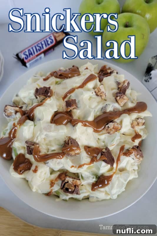 White bowl with snickers and apple salad covered in a bit of caramel, full size snickers bar and 3 green apples above the bowl.
