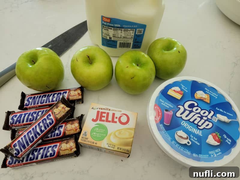 All the essential ingredients for Snickers Apple Salad laid out on a clean white counter: multiple full-size Snickers bars, a box of instant vanilla pudding, a container of Cool Whip, four crisp green apples, a milk jug, and a sharp knife, ready for preparation.