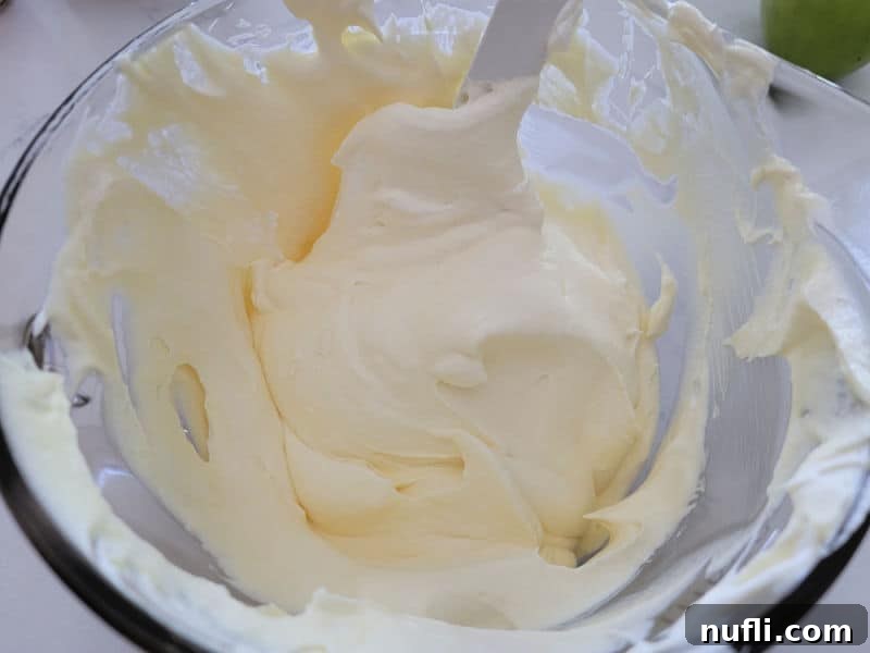 A clear glass bowl showcasing a creamy mixture of set vanilla pudding and folded-in Cool Whip, forming the light and airy base for the Snickers Apple Salad.