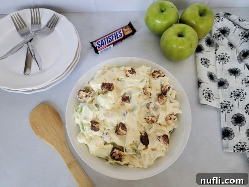 Snickers apple salad in a white bowl on a countertop with 3 apples, white plates with forks, and a Snickers Bar