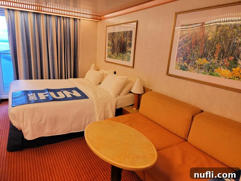 Carnival Stateroom with a oval table next to an orange couch, photo of flowers aboe the couch, bed with a blue Choose Fun mat over the top of it, paining print above the bed, blue and tan curtains next to a balcony door