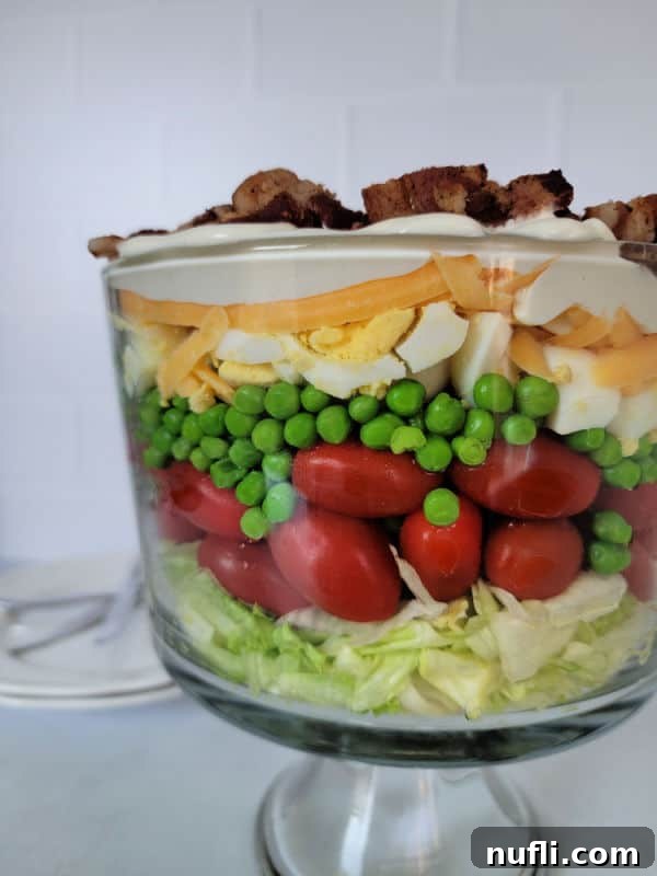 7 layer salad in a trifle dish with lettuce, tomato, peas, hard boiled eggs, dressing, cheese, and bacon. 