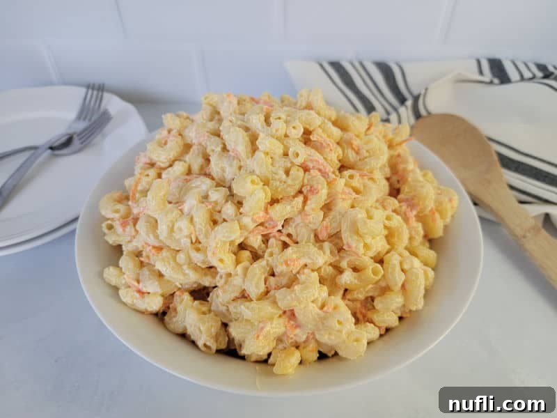 Macaroni salad with shredded carrots in a white bowl next to a wooden spoon, cloth napkin, and plates with forks. 