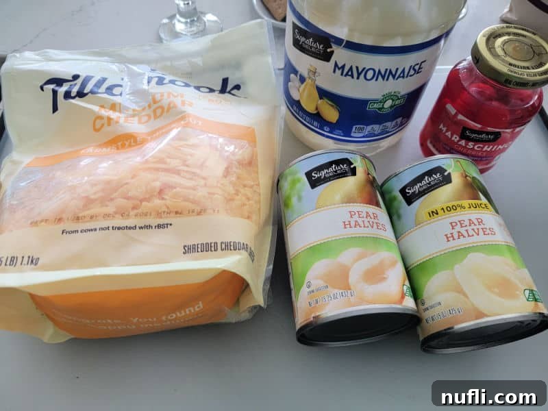 Close-up shot of the simple ingredients needed for Southern Pear Salad: a block of cheddar cheese, a jar of mayonnaise, maraschino cherries, and two cans of pear halves, all neatly arranged on a kitchen counter.
