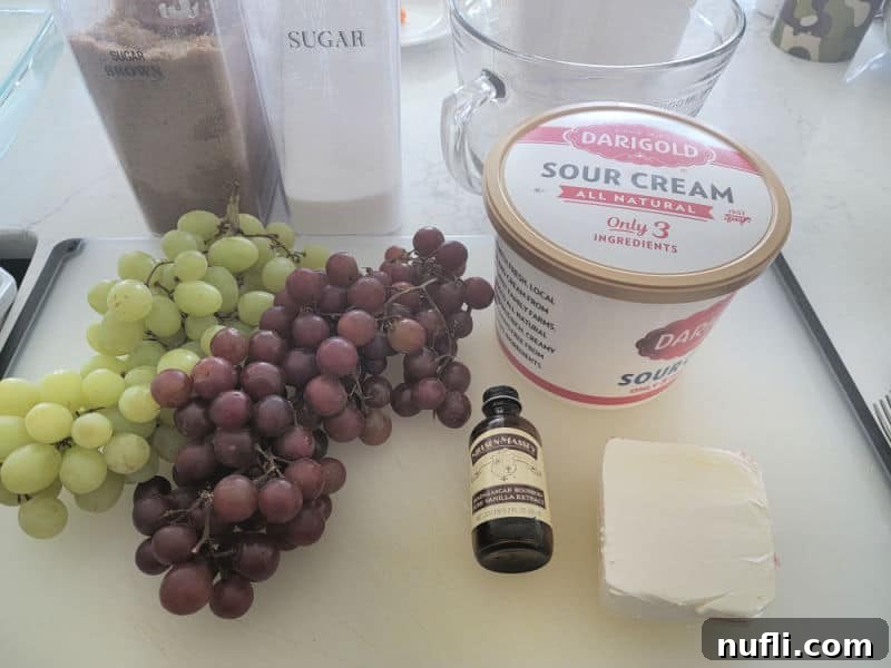 A collection of grape salad ingredients including brown sugar, granulated sugar, a glass bowl, sour cream, red and green grapes, vanilla extract, and cream cheese, neatly arranged on a kitchen counter.