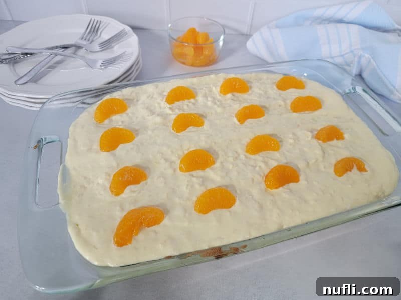 Pig Pickin Cake in a glass baking dish, next to a bowl of mandarin oranges, plates, and forks