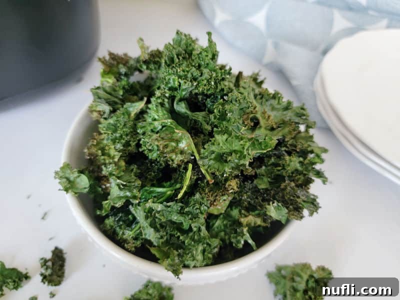 A bowl of crispy kale chips surrounded by plates and an air fryer, showcasing a delicious healthy snack.