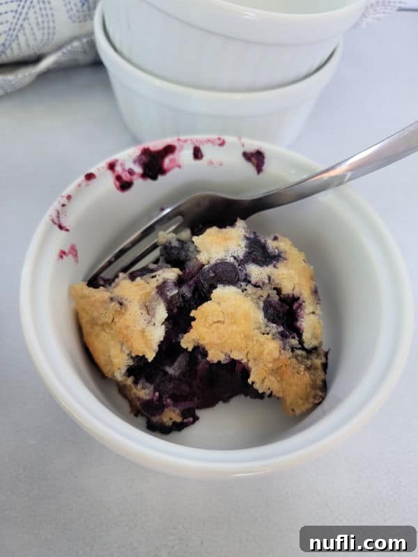 Small white bowl with blueberry cobbler and a fork next to a stack of bowls. 