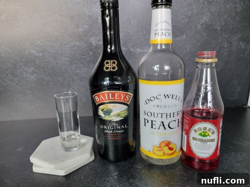 Brain Hemorrhage Shot Recipe Guide 3 A clear shot glass sits on two marble coasters, flanked by bottles of Baileys Irish Cream, Southern Comfort Peach Schnapps, and a bottle of Grenadine syrup, showcasing the essential ingredients.