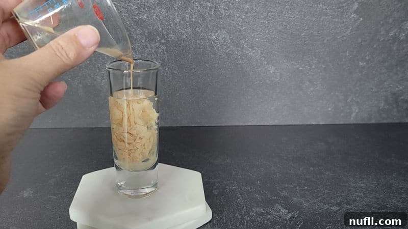 Brain Hemorrhage Shot Recipe Guide 5 Creamy white Baileys Irish Cream being gently poured over the clear peach schnapps in a glass shot glass, beginning to curdle and form brain-like patterns.