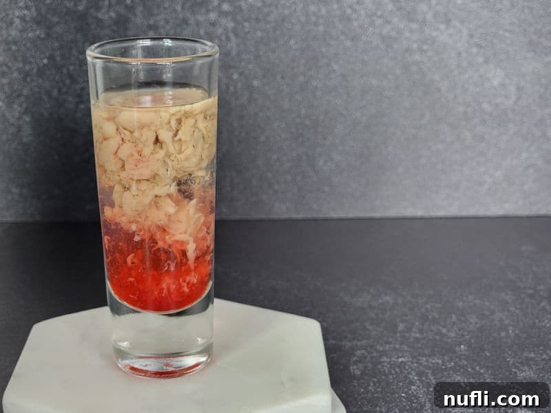 Brain Hemorrhage Shot Video Recipe 7 The mesmerizing swirling effect of cream and red liquid against a clear base in a Brain Hemorrhage Shot, presented in a shot glass on a marble coaster.