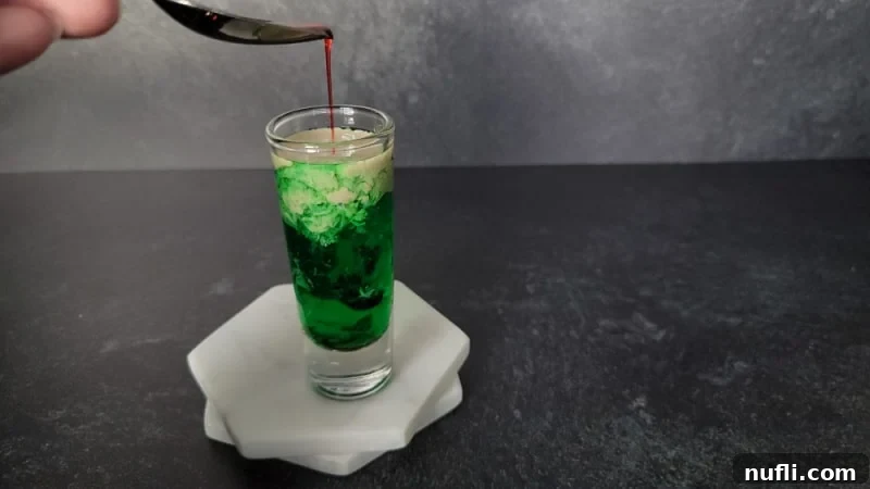 Spoon above a shot glass with red liquid dripping off of it into a green and cream liquid shot