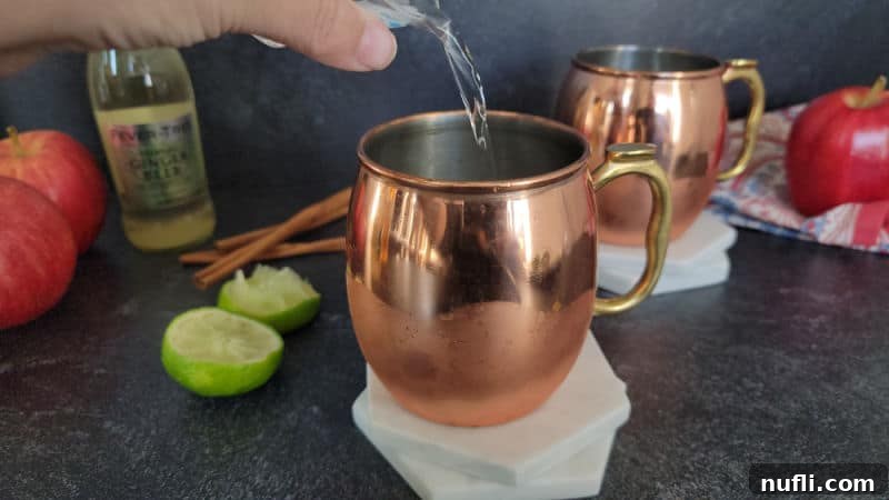 Apple Cider Mule Cocktail 4 Clear liquid pouring into a copper mule mug on a coaster with apples, lime pieces, and ginger beer in the background.