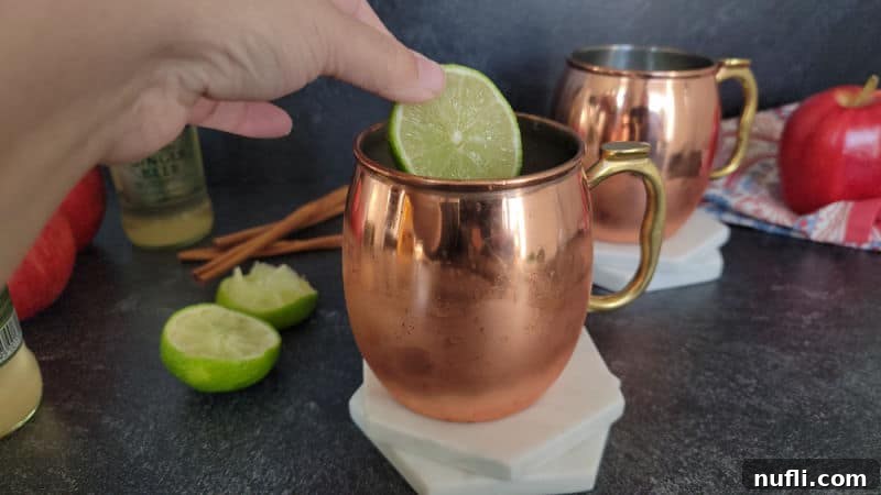 Apple Cider Mule Cocktail 5 lime wheel being placed in a copper mule mug on two coasters, additional mug, apples and limes in the background