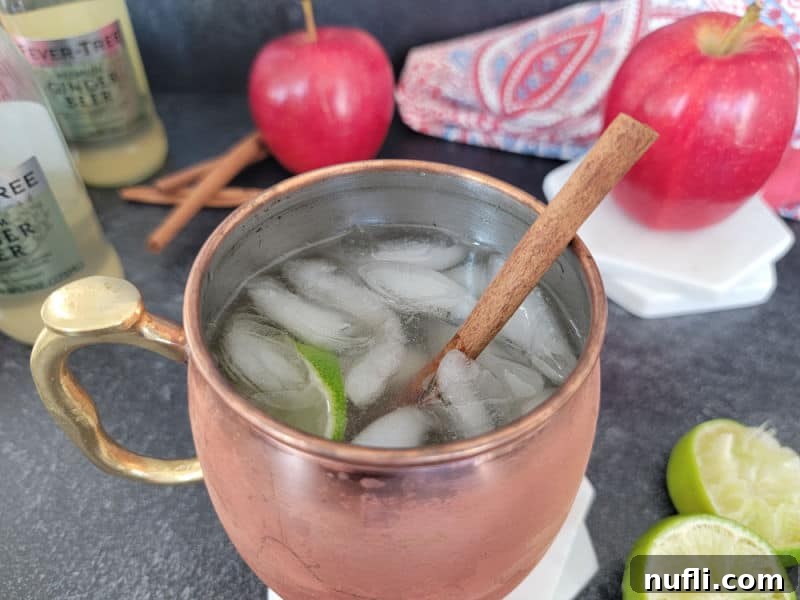 Apple Cider Mule Cocktail 6 Cinnamon stick in a mule mug with liquid and ice. Next to lime wheels, apples, and ginger beer