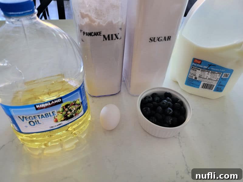Ingredients for Bisquick Blueberry Muffins laid out: vegetable oil, pancake mix, sugar, egg, blueberries, and milk on a white counter