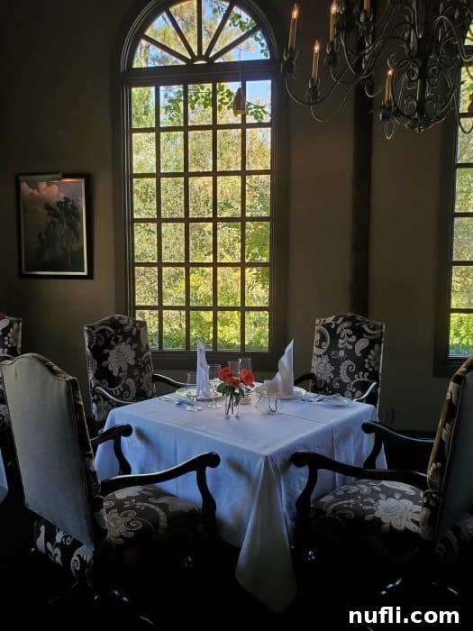 Table covered in white linen tablecloth, and place settings next to fancy chairs with a chandelier above. 