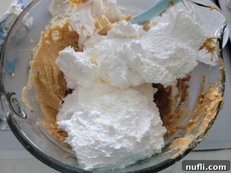 A generous dollop of Cool Whip delicately placed on top of the mixed pumpkin dip base in a glass batter bowl, ready to be folded in for a fluffy texture.