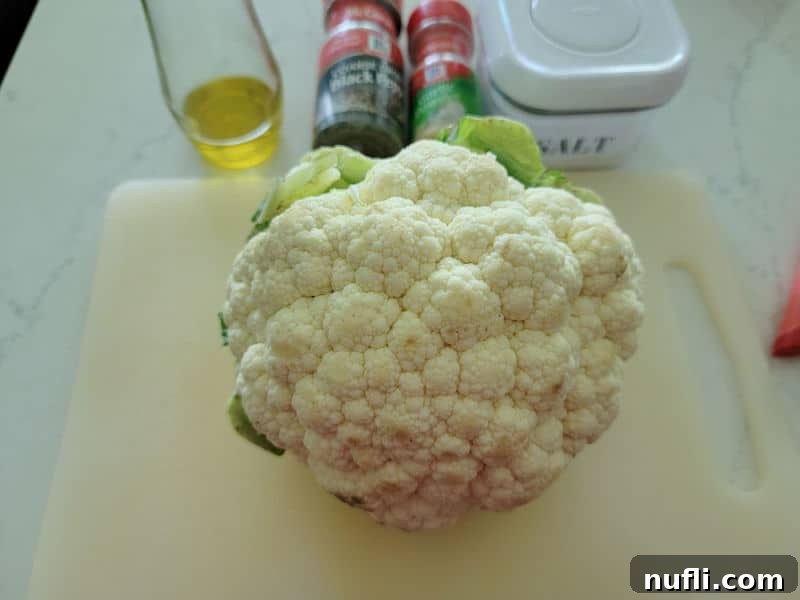 A pristine white cutting board holds a fresh head of cauliflower, surrounded by small bowls of salt, black pepper, and aromatic garlic powder, ready for seasoning.