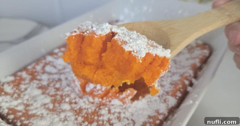 Easy Fluffy Carrot Soufflé 11 A generous serving of carrot soufflé on a wooden spoon, held above a white casserole dish, highlighting its creamy texture and powdered sugar topping.