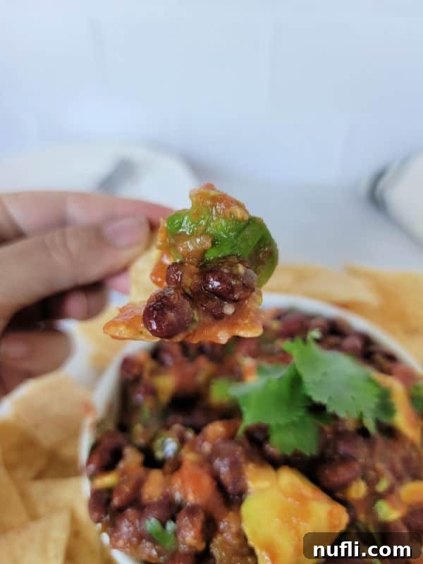 A hand holding a crunchy tortilla chip generously loaded with vibrant black bean salsa, poised over a bowl of more salsa and chips.