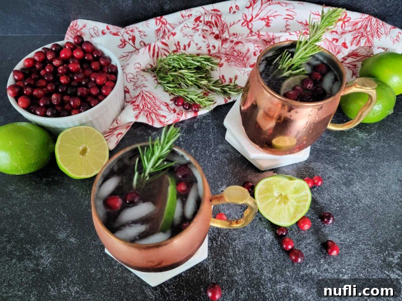 white bowl filled with cranberries next to a red and white cloth napkin, limes, rosemary, and two copper mule mugs with cranberry moscow mules in them