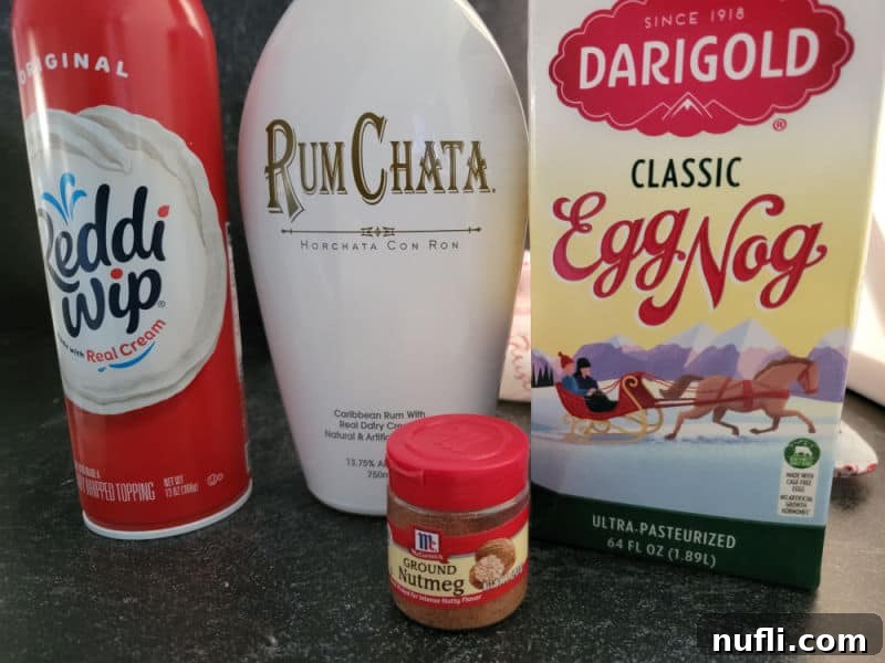Ingredients for a RumChata Eggnog Cocktail laid out on a kitchen counter: a can of whipped cream, a bottle of RumChata, a carton of eggnog, and a container of ground cinnamon, ready for mixing.