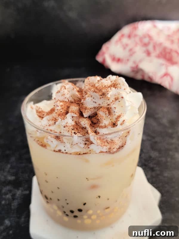 A beautifully prepared Eggnog drink in a glass, topped with a generous dollop of whipped cream and a sprinkle of nutmeg. A soft cloth napkin peeks from the background, adding a touch of elegance.