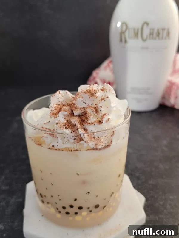 A bottle of RumChata standing behind a glass filled with creamy eggnog, highlighting the two main ingredients of the cocktail.