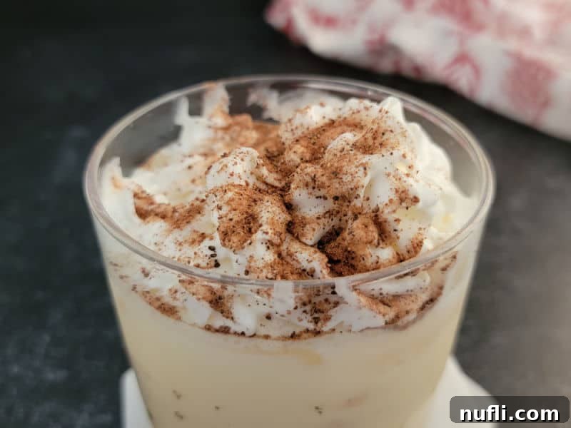 Close-up of an eggnog drink crowned with whipped cream and freshly ground nutmeg, with a soft cloth napkin blurred in the background, emphasizing the luxurious texture and festive garnish.