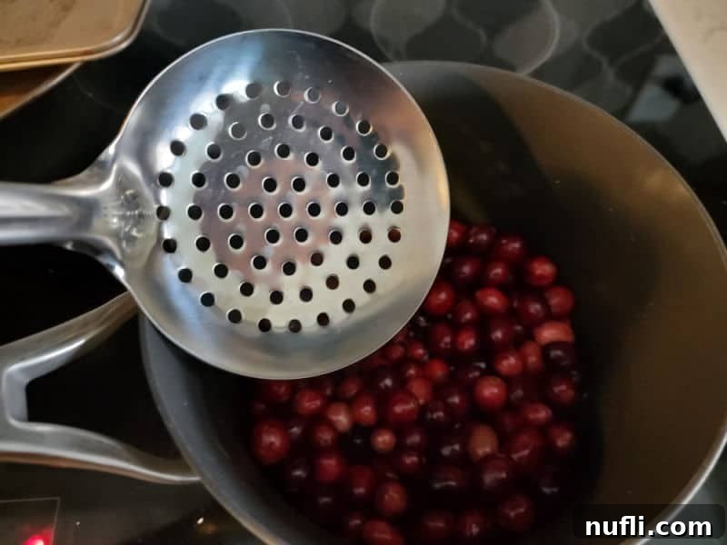A slotted spoon carefully removing simple syrup-coated cranberries from a saucepan