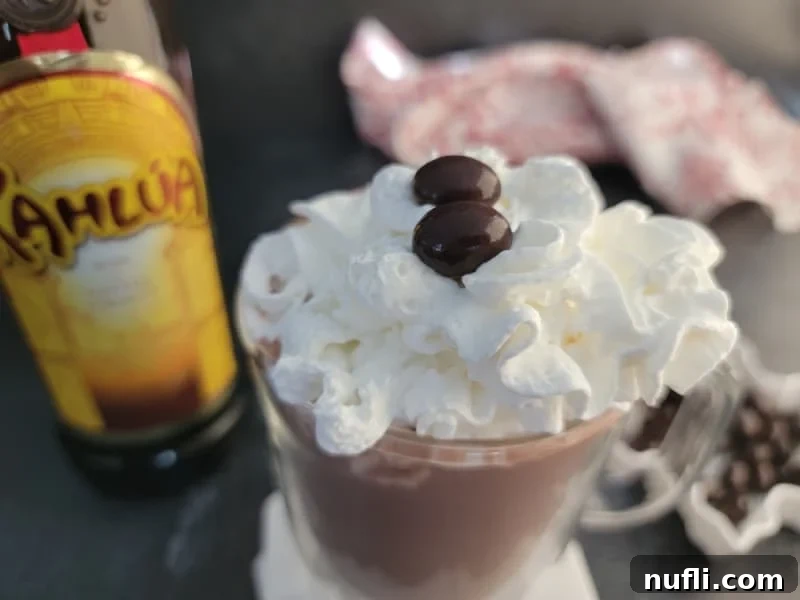 Decadent Kahlua Hot Chocolate 4 A glass mug filled with Kahlúa Hot Chocolate, generously topped with whipped cream, sits beside a bottle of Kahlúa, highlighting the key ingredient.