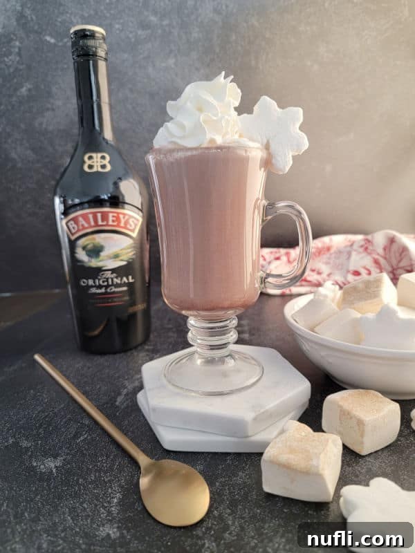 Effortless Baileys Hot Chocolate 4 Baileys Hot Chocolate in a glass mug with whipped cream next to a bottle of Original Baileys Irish Cream, a bowl of marshmallows, a gold spoon, with a cloth napkin